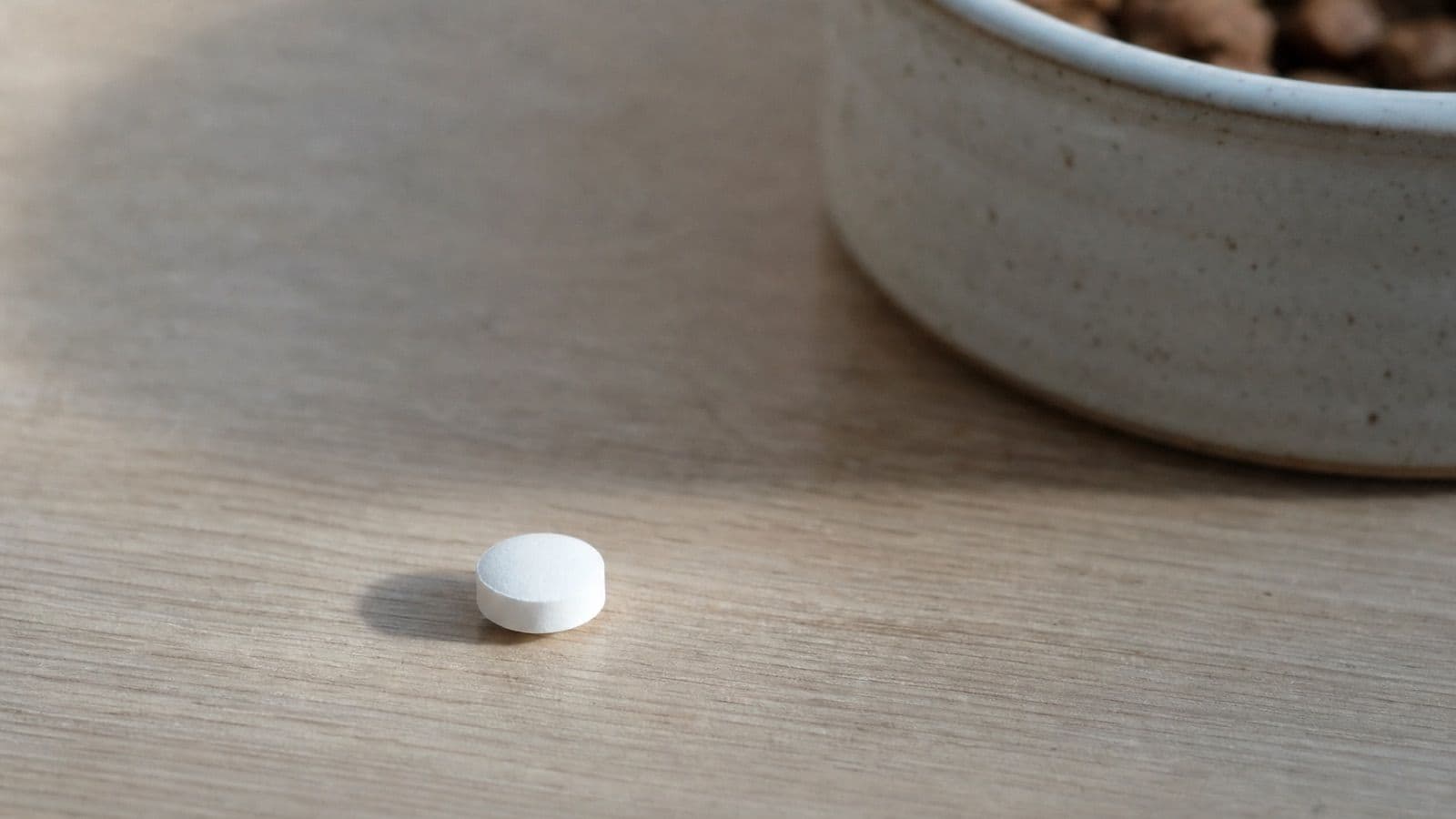 A single small white tablet on a pale oak surface beside a ceramic bowl of food